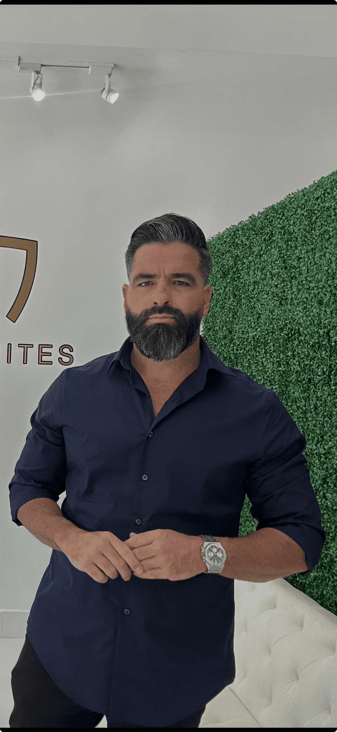 Man with a salt-and-pepper beard in a navy shirt against a green hedge wall.